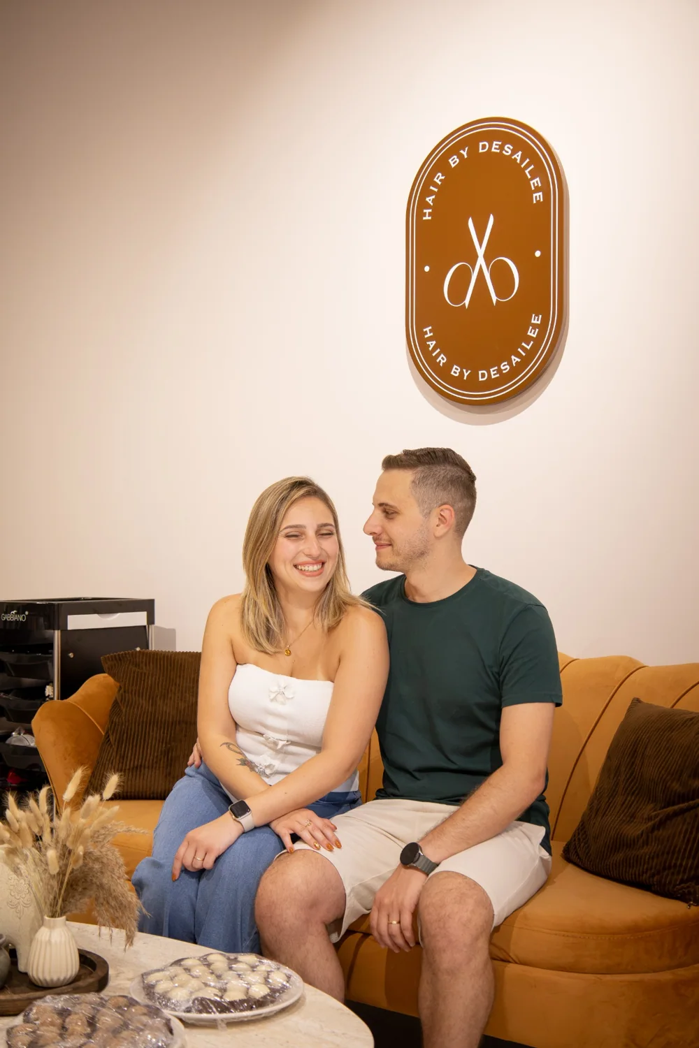 Desailee and her husband seated together beneath the salon sign